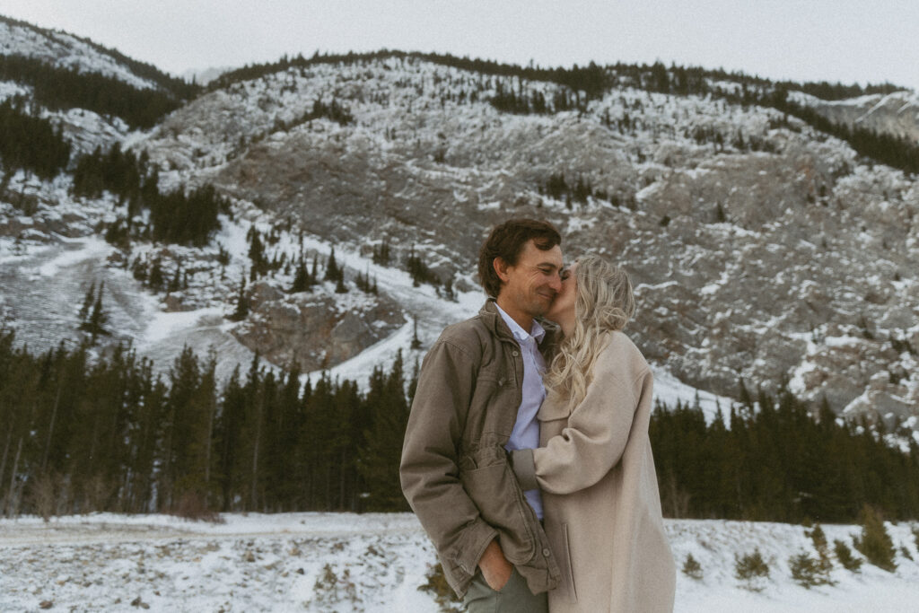 young couple holds each other wearing jackets in front of a mountain in the winter