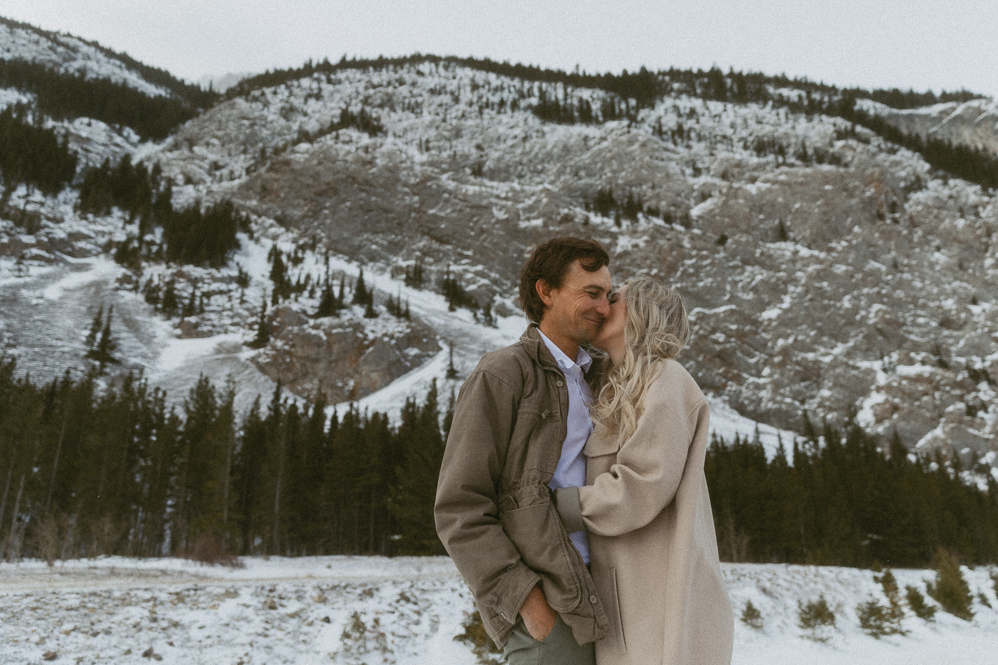 Man and woman snuggle up to each other with a mountain behind them in the winter