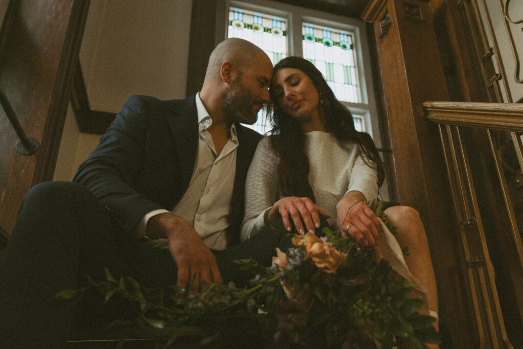 Bride and groom sit on stairs at the courthouse with bouquet in hand resting heads against each other