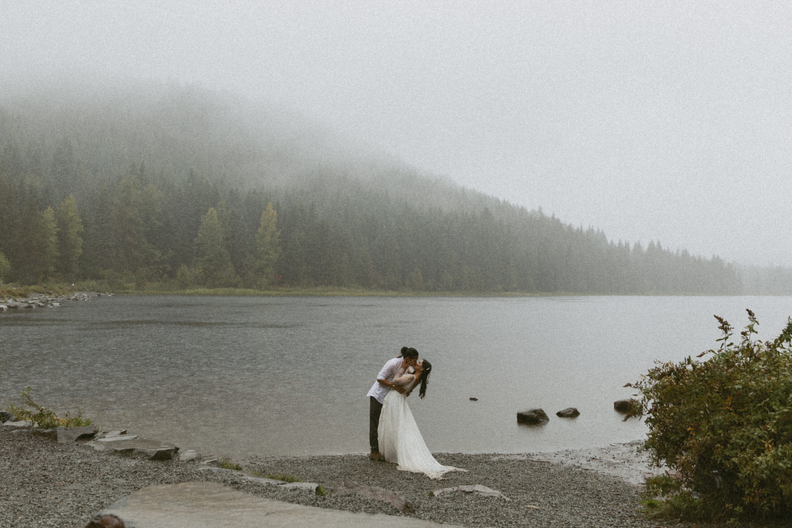 Bridge and groom dip kiss in front of a lake on a rainy, moody day