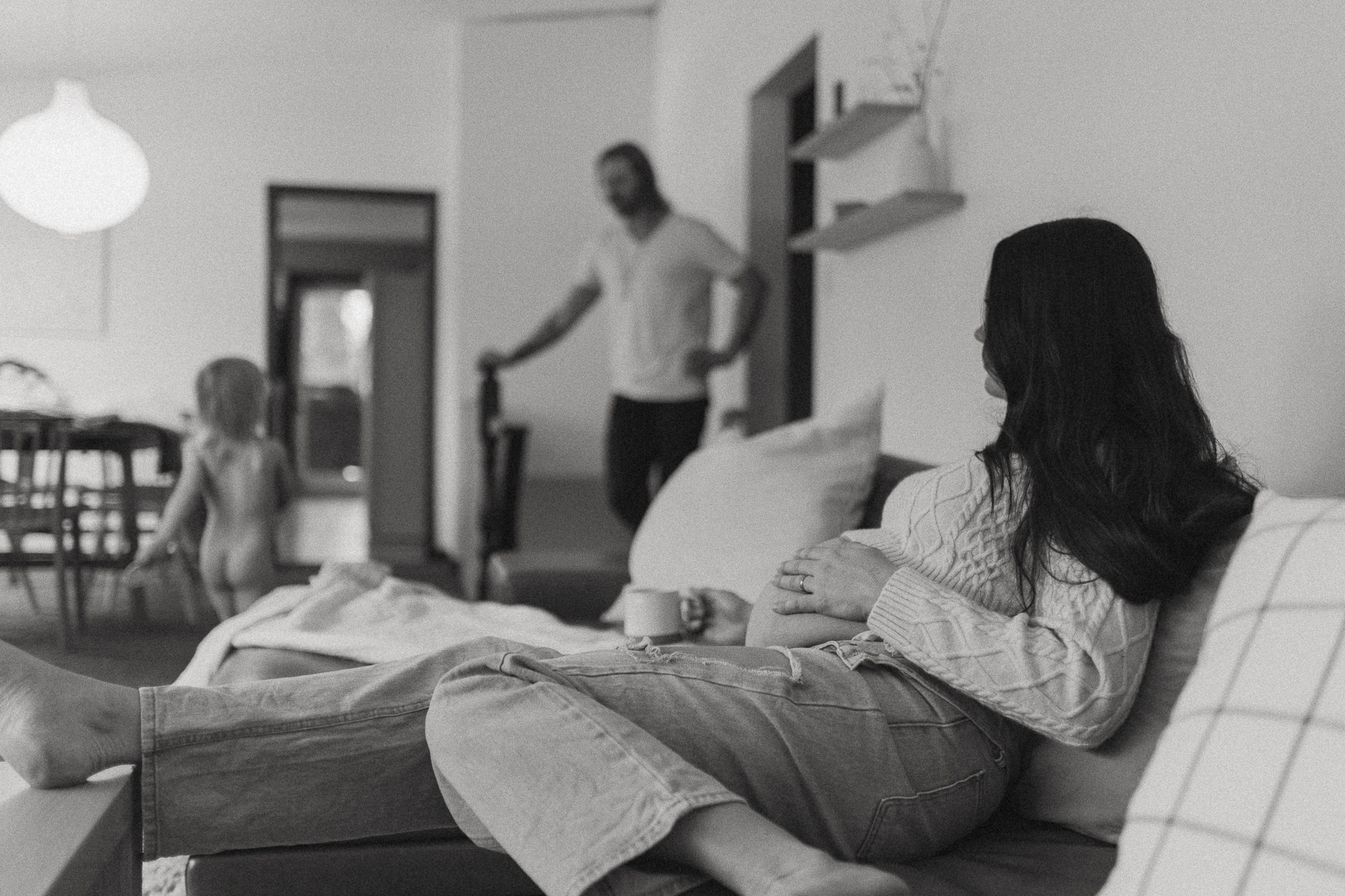 pregnant woman sits on couch while watching her son and husband in their living room