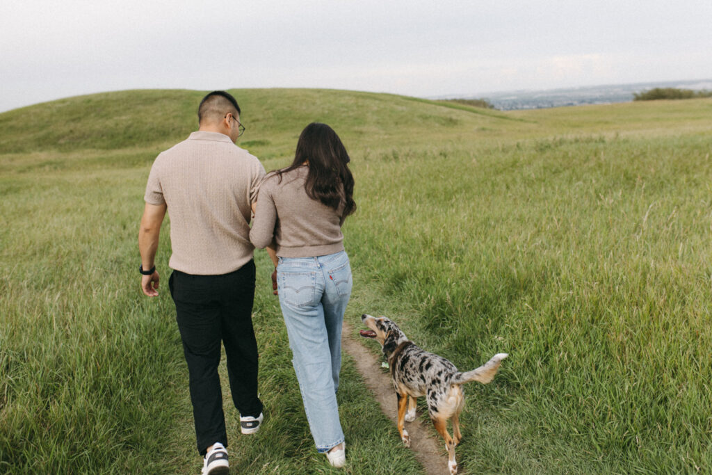 Couple walking hand-in-hand across Nose Hill Park with Calgary skyline in the background, Alberta engagement photographer capturing natural moments.